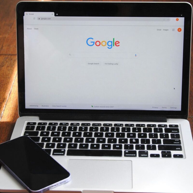 open laptop on wooden surface with cell phone resting on keyboard. Google search engine window pulled up on screen